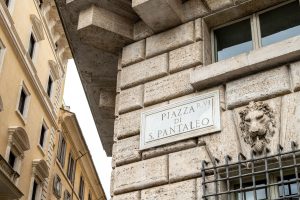 Targa stradale in marmo di Piazza di S. Pantaleo accanto a una testa di leone scolpita su un edificio storico.