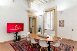 Sala riunioni con tavolo ovale, sedie bianche, soffitto a cassettoni e schermo con logo European School of Economics.
