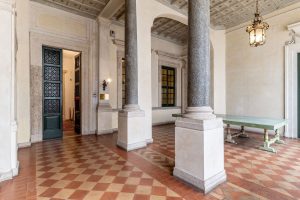 Soportal histórico con grandes columnas de piedra, suelo de terracota a cuadros, techo decorado y una larga mesa.