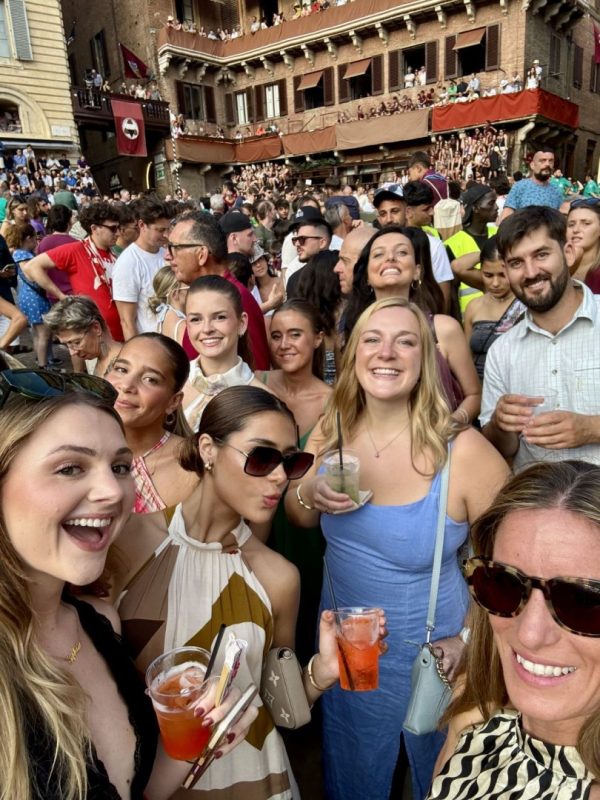 Un gruppo di studenti internazionali dell'ESE Florence scatta un selfie sorridente con dei drink in mano in Piazza del Campo, circondati dalla folla festante e dagli edifici storici durante il Palio di Siena.