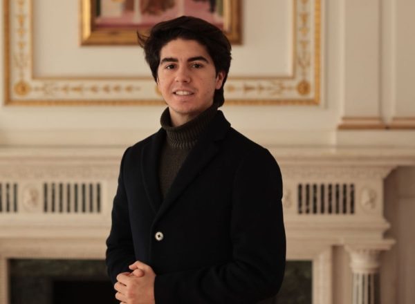 Retrato de Mattia de pie cerca de una chimenea decorada, con abrigo y jersey de cuello alto.