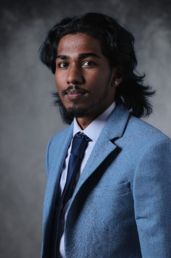 Retrato de Rudi con chaqueta de traje azul claro y corbata oscura sobre un fondo gris.